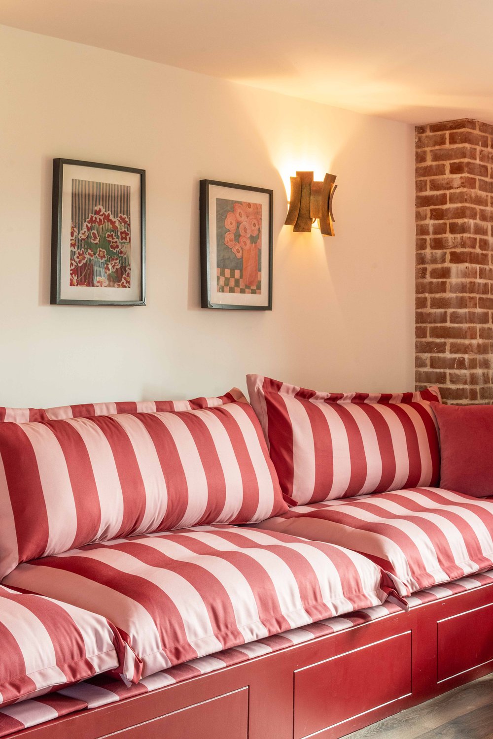 Red and pink candy-stripe daybed under two framed botanical prints and a brass sconce, against an exposed brick column