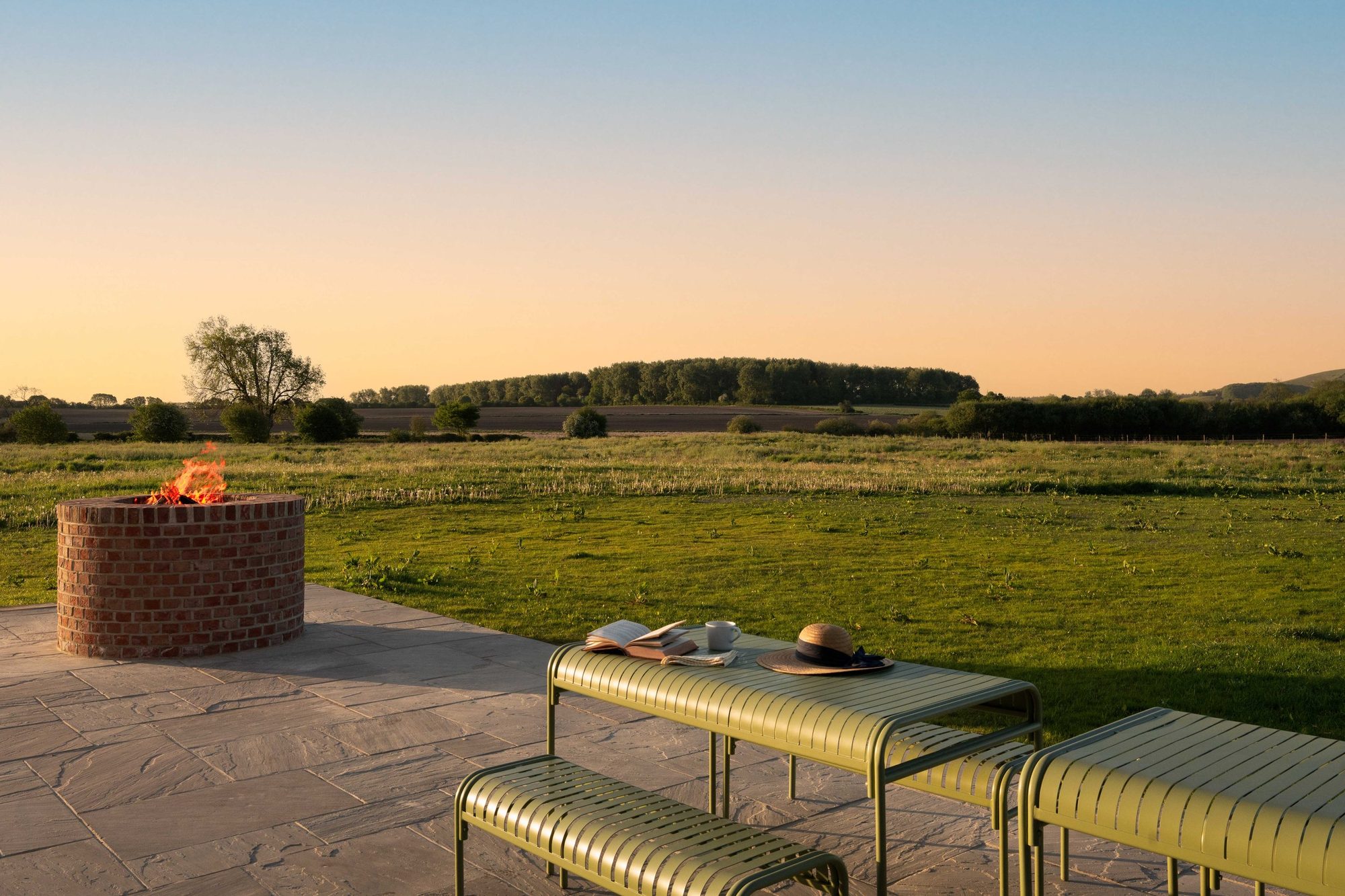 Round brick fire pit lit on a flagstone terrace with olive-green slatted benches, a hat and an open book overlooking fields