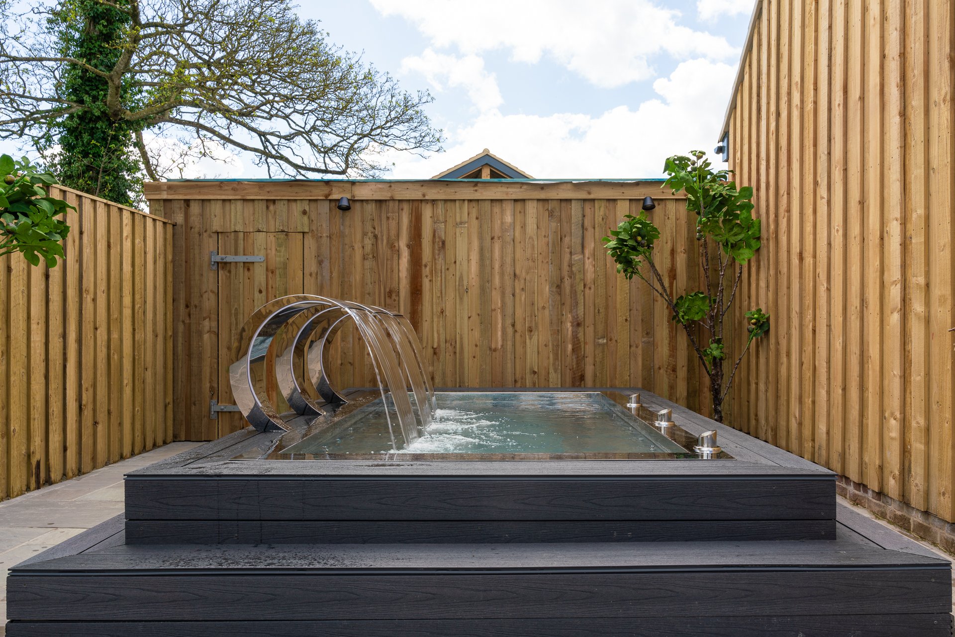 Square stainless-steel plunge pool with a mirrored waterfall arch on a black deck, against timber fencing with a young fig tree beside it