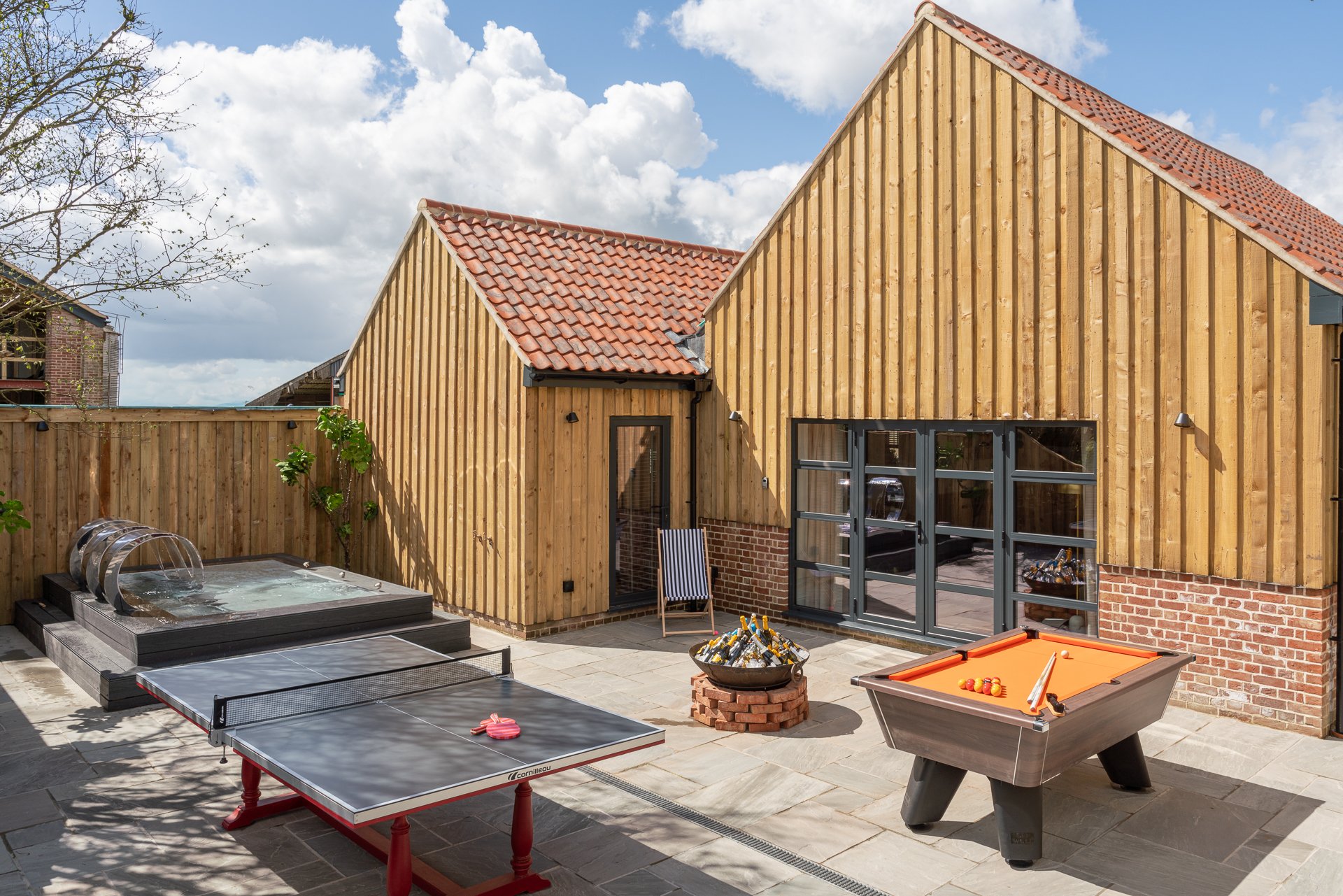 The Merchant's private courtyard between two timber-clad barns, with an XL stainless-steel hot tub, outdoor ping pong table, orange-topped pool table and a fire pit stacked with bottled beers