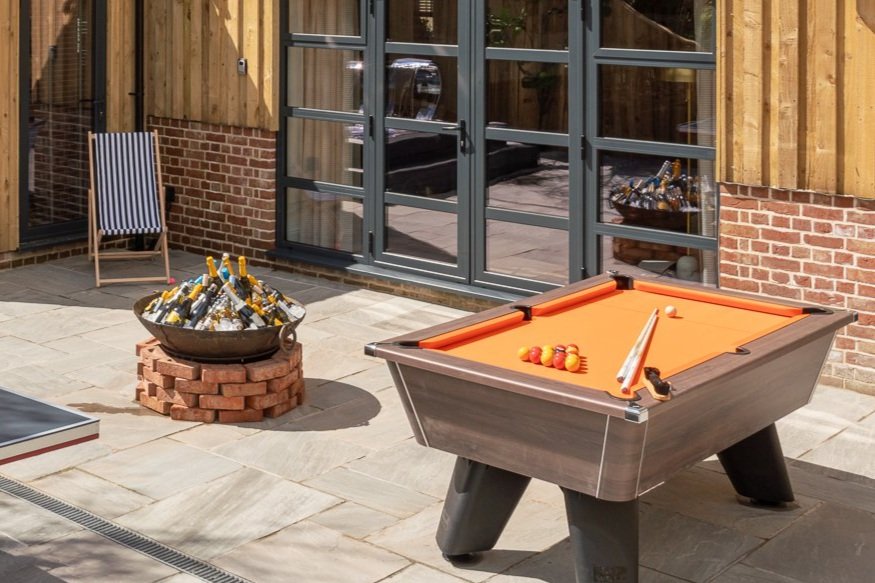 Orange-topped pool table on the patio beside a fire pit filled with bottled beers on ice and a navy striped deck chair