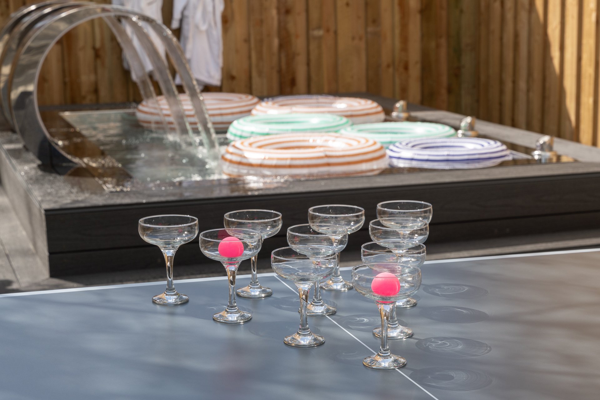 Coupe glasses lined up for prosecco pong on the outdoor table tennis table, striped ring floats bobbing in the hot tub behind