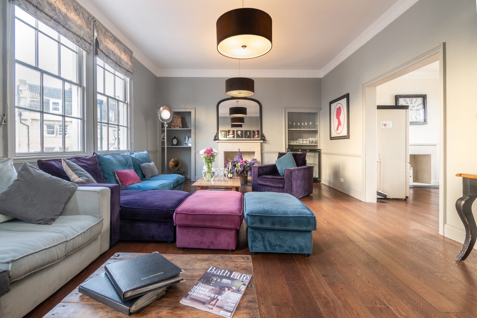 Lounge with grey, purple and teal velvet sofas, pink and blue ottomans and a drinks bar by tall sash windows