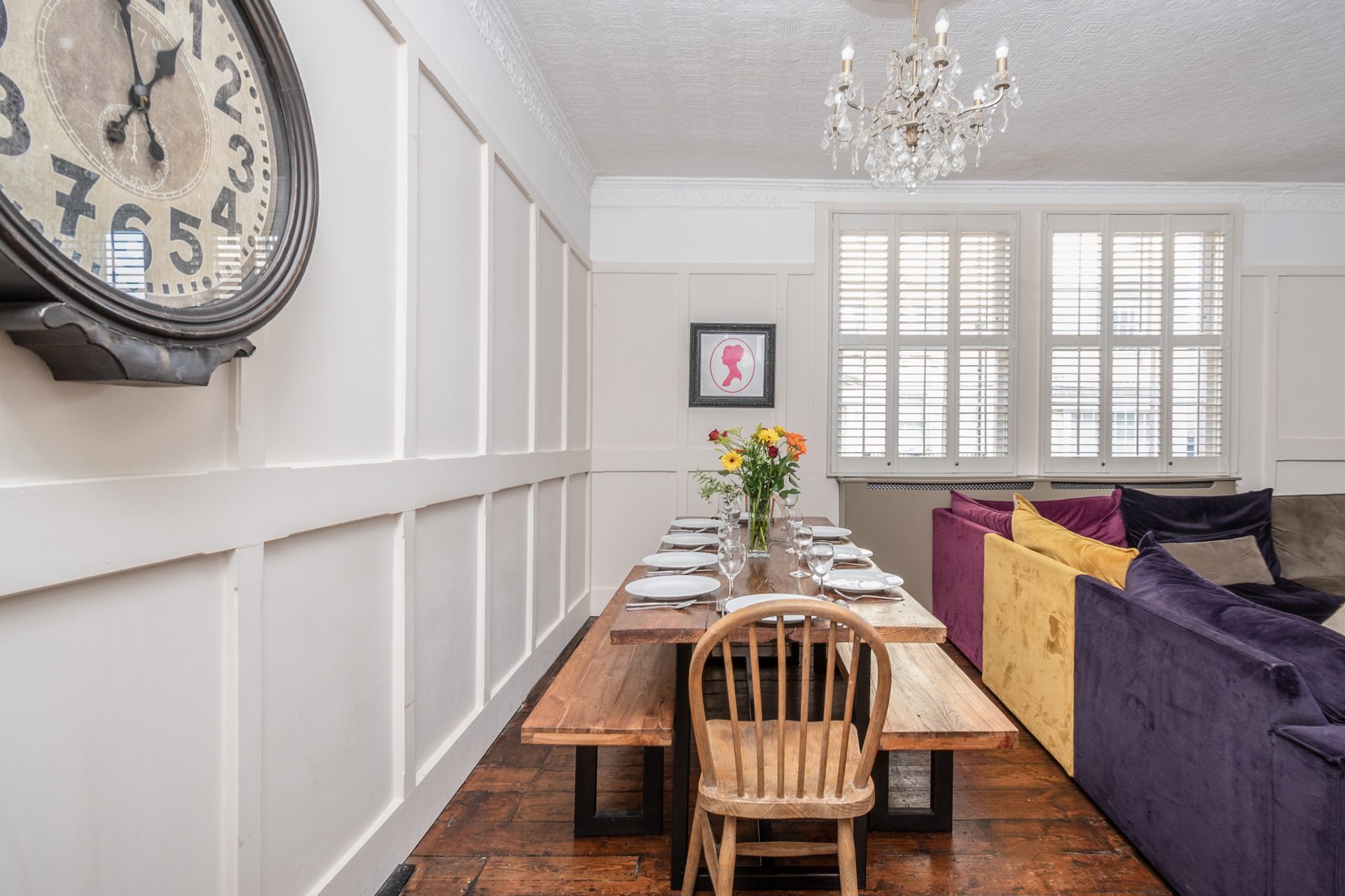 Dining room with long plank table, purple and yellow velvet sofa, crystal chandelier and oversized station clock