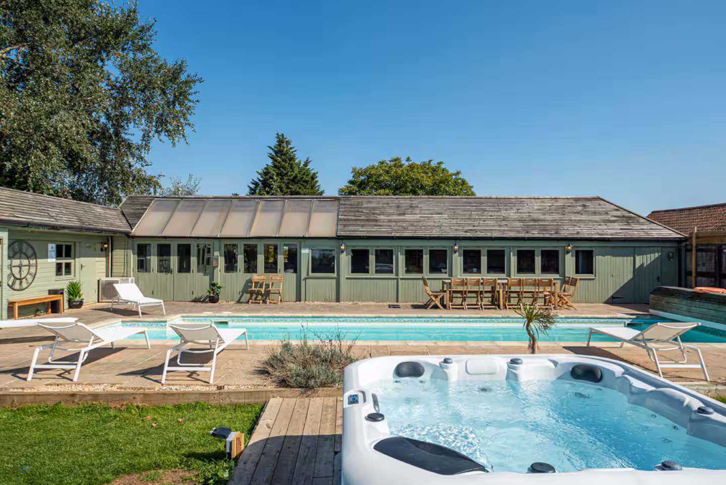Outdoor hot tub in the foreground with sun loungers and the heated pool stretching along the pool house behind