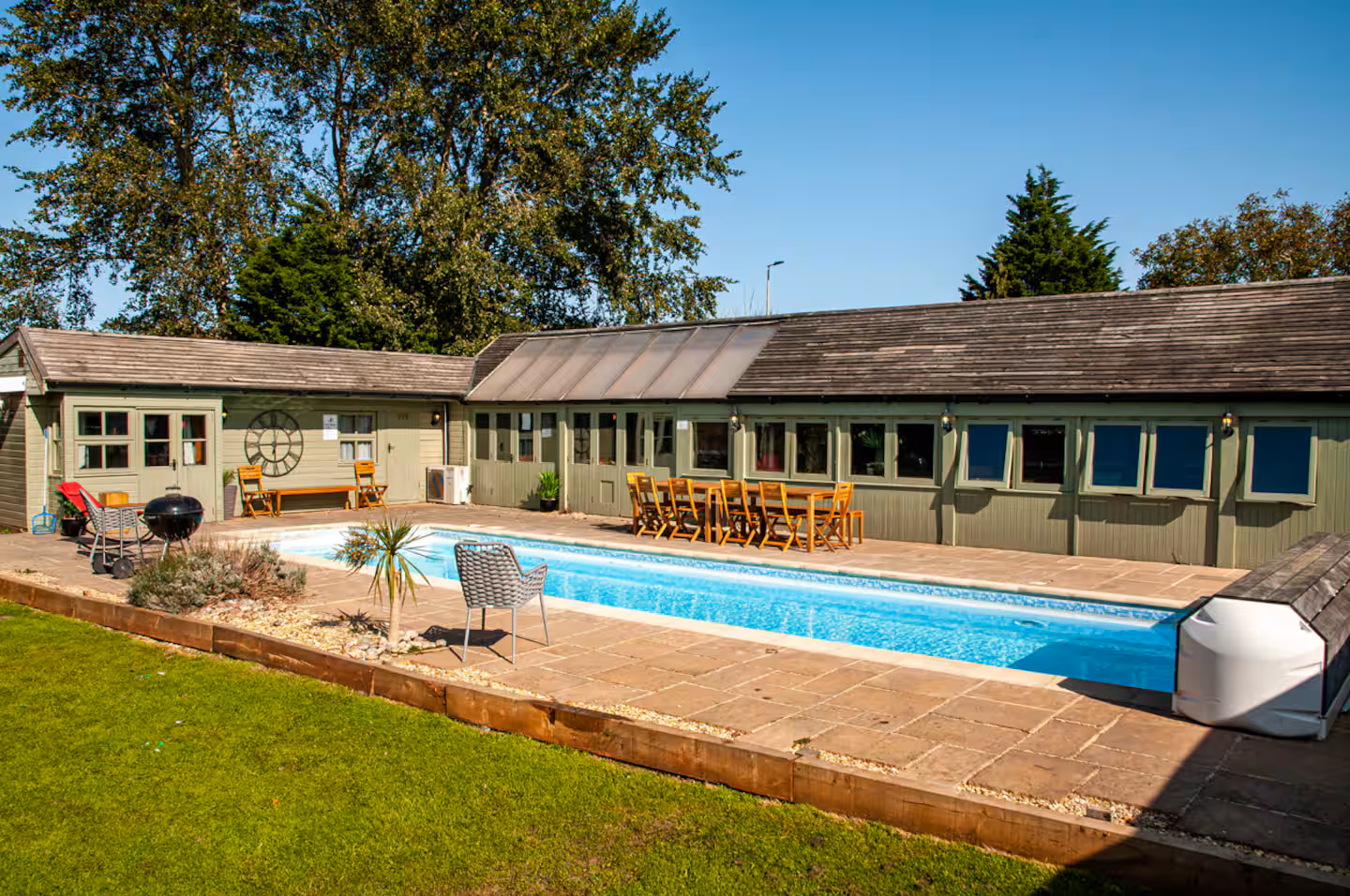 Wide angle of the pool with a row of dining chairs lined up along the pool house and BBQ to one side
