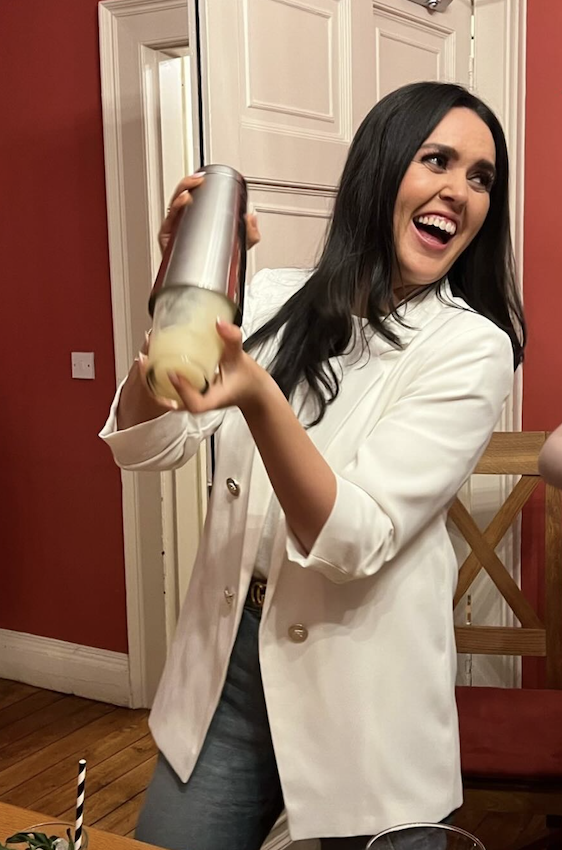 Hen shaking a cocktail in a white blazer with cocktail glasses on the table